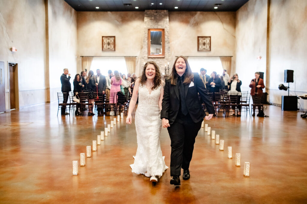 Two people joyfully walk hand in hand down a candle-lit aisle, with guests smiling in the background.
