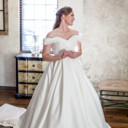 Bride in an off-the-shoulder white gown stands smiling in a sunlit room with wooden floors.