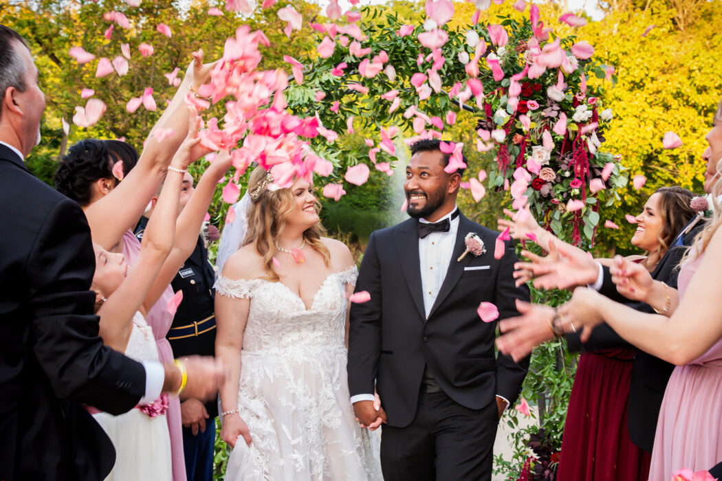 Bride and groom holding hands, smiling as guests throw pink flower petals in celebration.