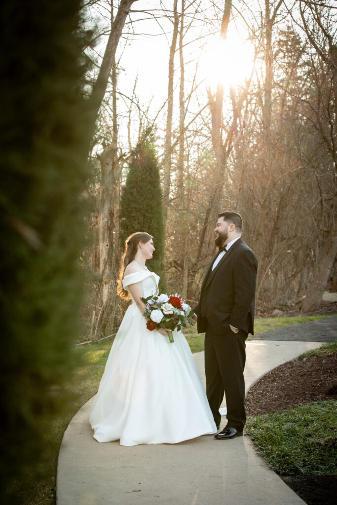 Bride and groom standing on a garden path, smiling at each other, with sunlit trees in the background.