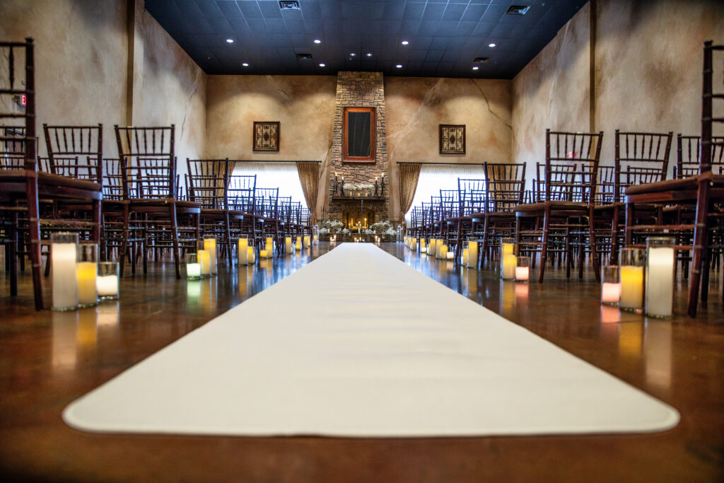 A wedding aisle with candles on the floor is lined with empty chairs, leading to a fireplace in a dimly lit room.