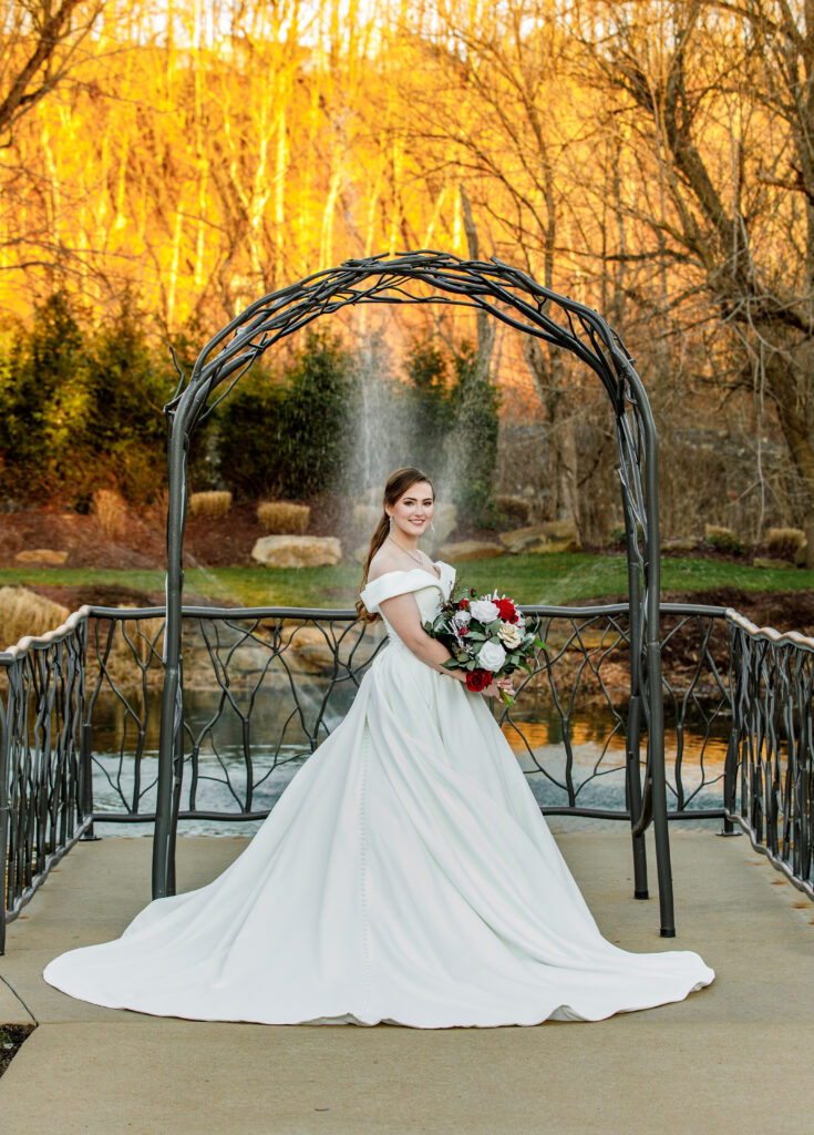 Bride in a white gown holding a bouquet, standing under an arch near a pond with autumn trees in the background.