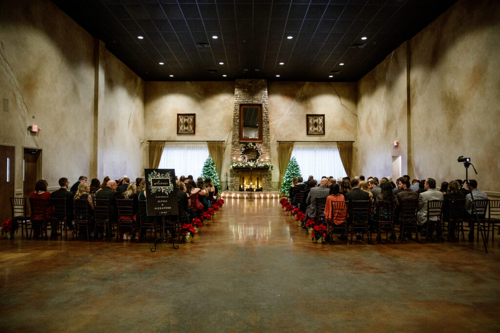 Indoor wedding ceremony with guests seated, facing a fireplace adorned with greenery and decorations.