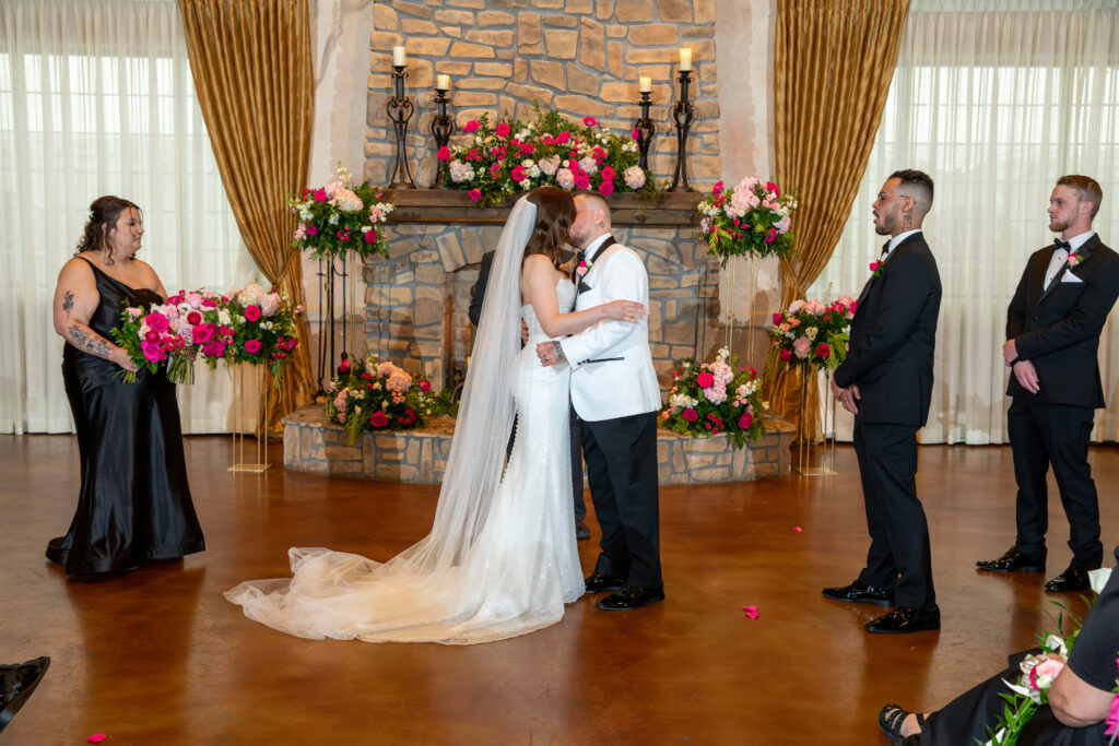 Bride and groom kiss at a wedding ceremony, surrounded by wedding party and floral decorations.