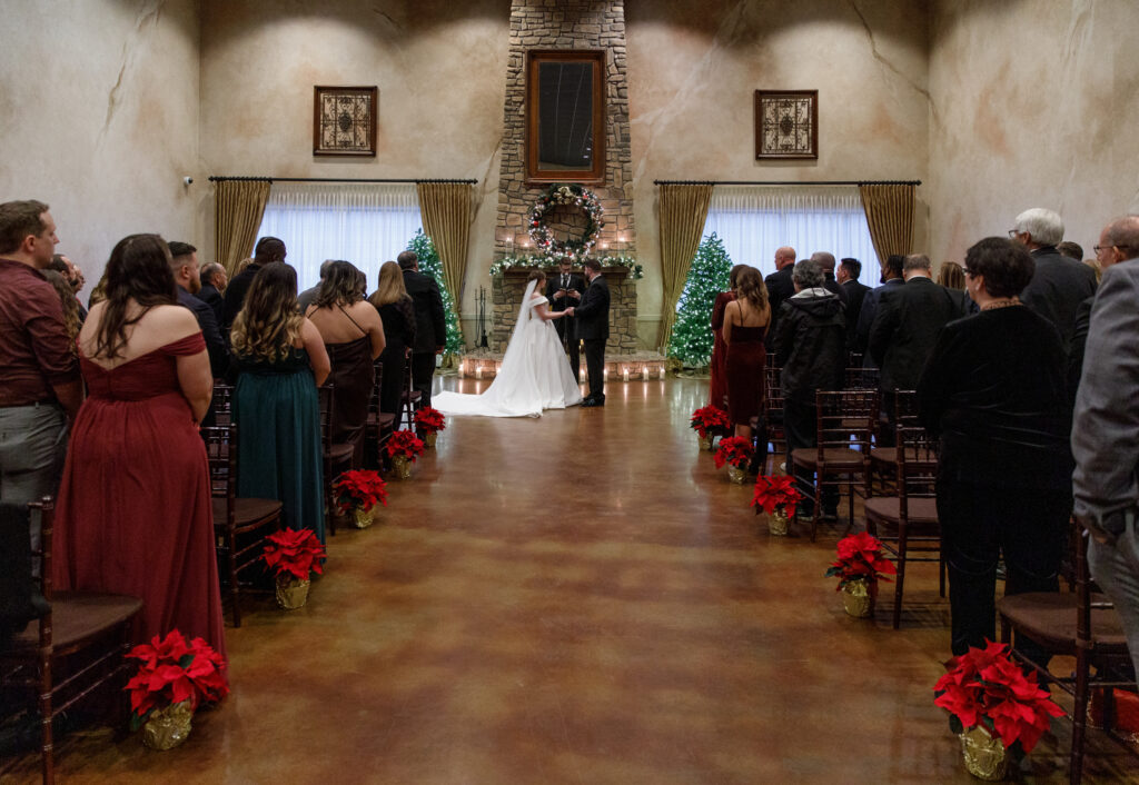 A wedding ceremony in a decorated hall with a couple holding hands, surrounded by guests, red flowers, and Christmas trees.