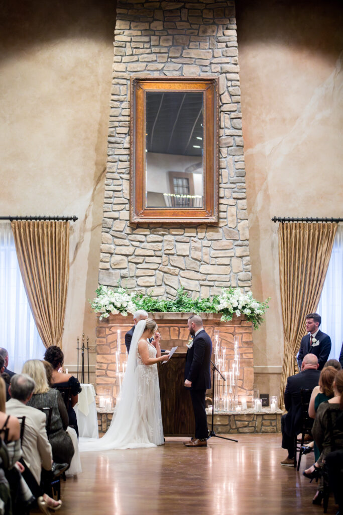 Bride and groom standing at an indoor altar with a stone backdrop, surrounded by guests.