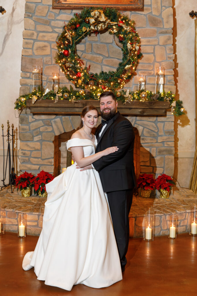 A couple in formal attire poses in front of a decorated stone fireplace, with a festive wreath and candles.