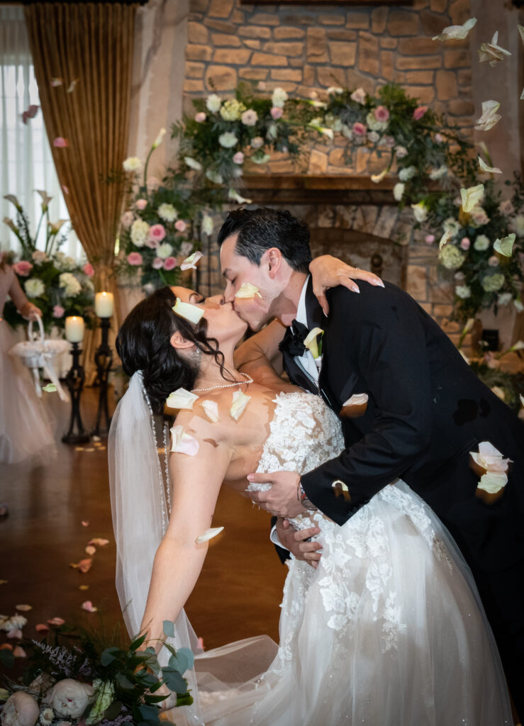 A couple kisses in a wedding venue decorated with flowers, as rose petals fall around them.