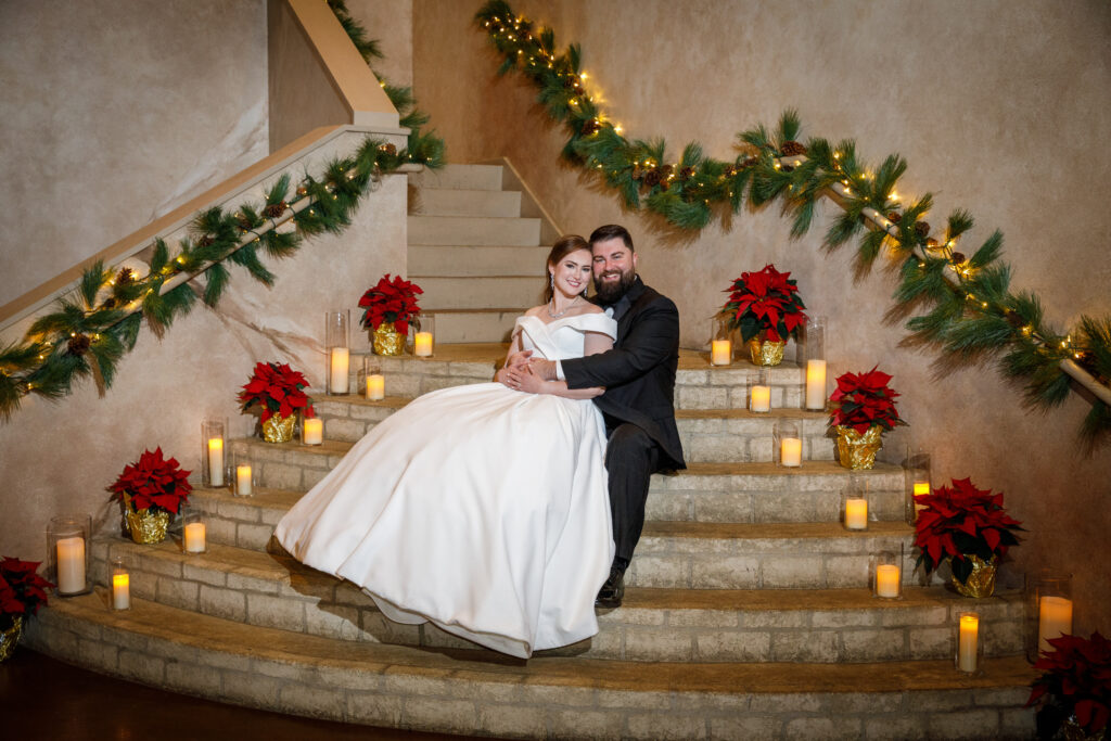 Bride and groom sitting on decorated stairs with poinsettias and candles, smiling.