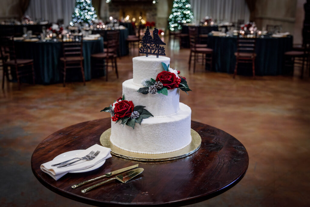 A three-tier white wedding cake with red roses on a wooden table, set in a dimly lit banquet hall with Christmas decor.