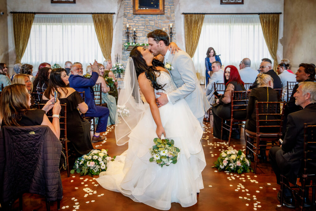 Bride and groom kiss in the aisle while guests watch during a wedding ceremony in a decorated venue.