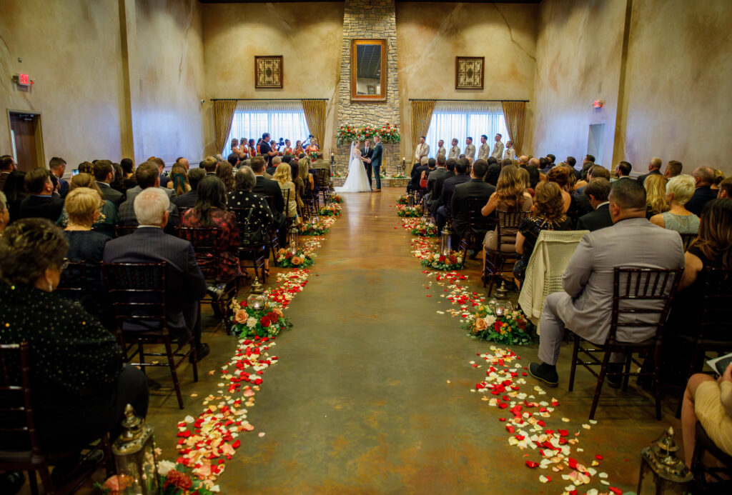 A wedding ceremony in a hall with guests seated, floral decorations, and a couple at the altar.