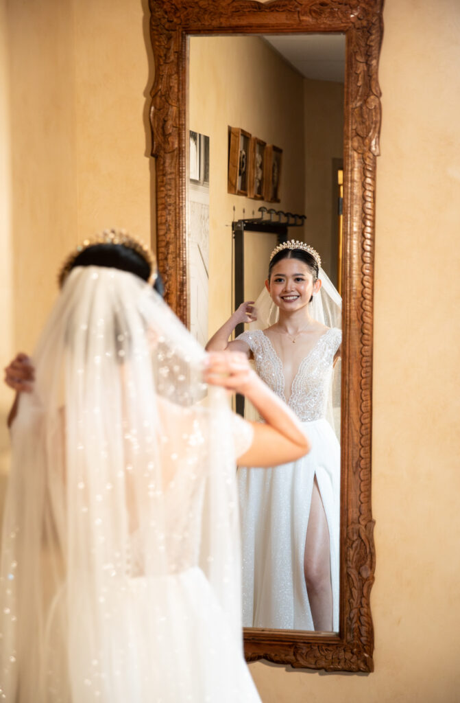 A bride adjusts her veil in front of an ornate mirror, smiling in a room with warm-toned walls.