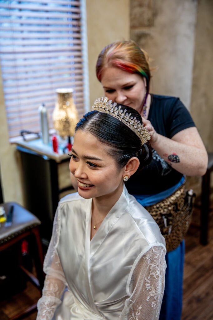 A smiling bride in a robe gets her hair styled by a hairstylist, wearing a tiara, in a room with wooden blinds.