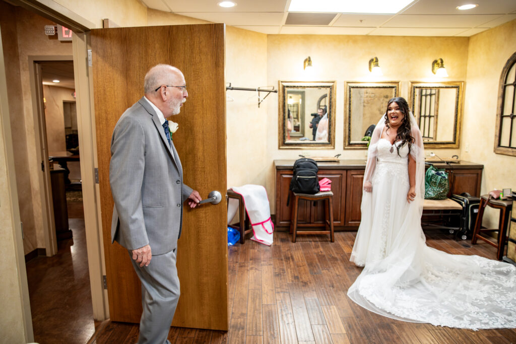 An elderly man in a suit enters a room to see a smiling bride in her wedding dress.