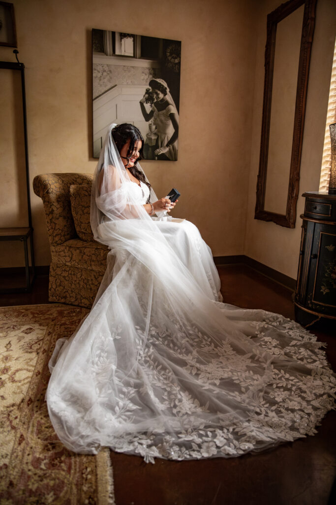 Bride in white gown sits on a chair, looking at her phone. A black-and-white portrait is on the wall behind her.