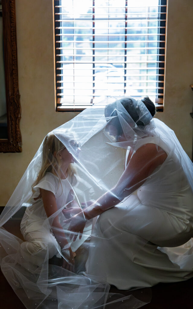 Bride and young girl smiling under a veil in front of a window with wooden blinds.