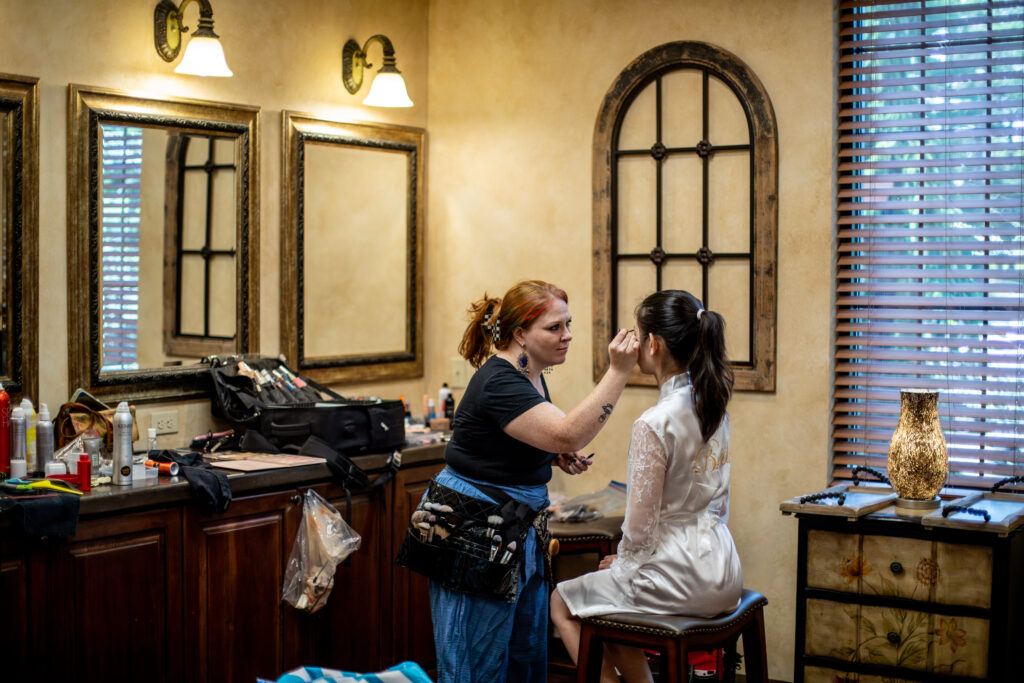 Makeup artist applying makeup to a seated woman in a salon with mirrors and warm lighting.