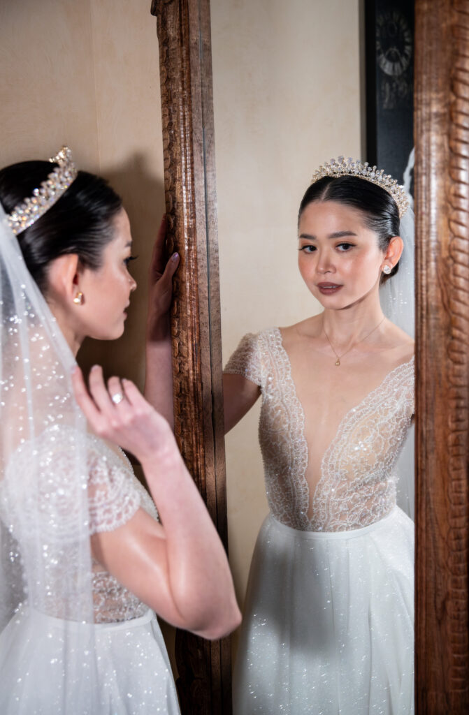 Bride in sparkling dress and tiara looks at herself in a mirror with a soft expression.