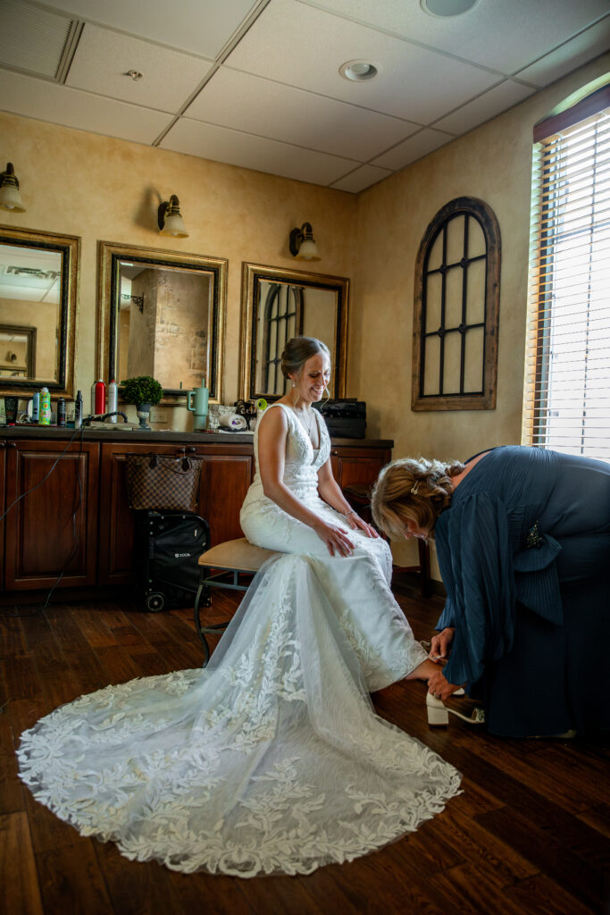 Bride in a white dress sits while another person helps adjust her shoe in a warmly lit room with mirrors and windows.