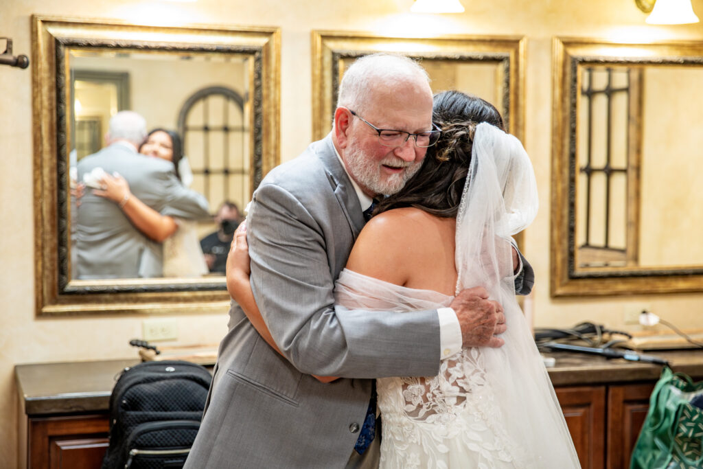 A bride in a lace dress embraces an older man in a suit, reflecting a joyful moment in a warmly lit room.