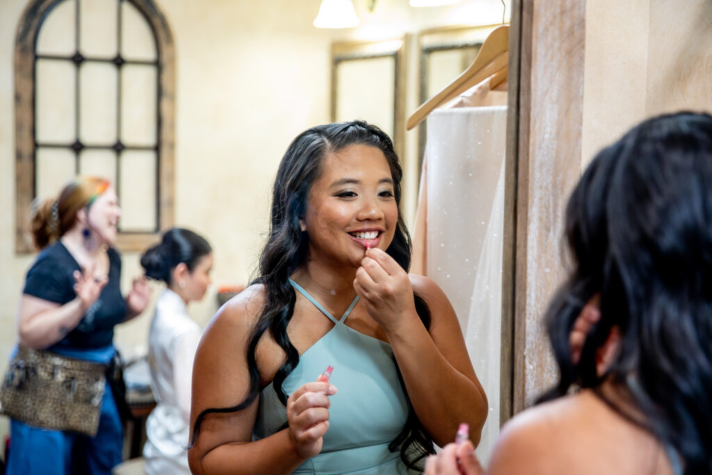 A woman in a blue dress is applying lipstick while looking in a mirror. Two others are in the background.