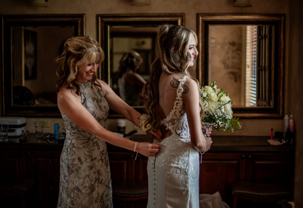 A woman helps a bride with her dress in a warmly lit room, surrounded by mirrors and flowers.
