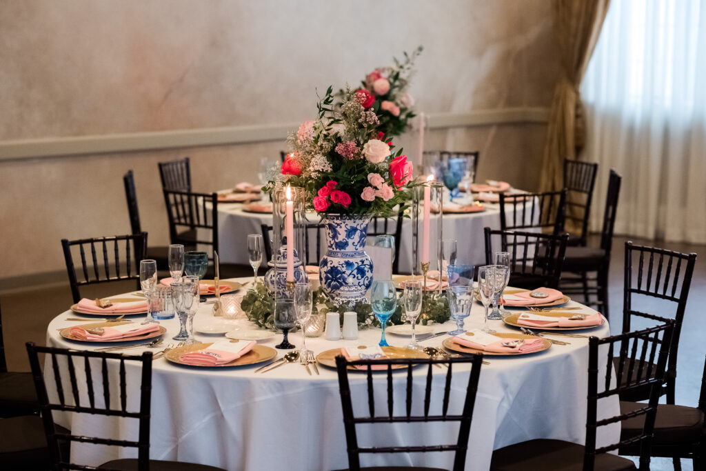Elegant table setting with floral centerpiece, blue and white vases, candles, and pink napkins on white tablecloths.