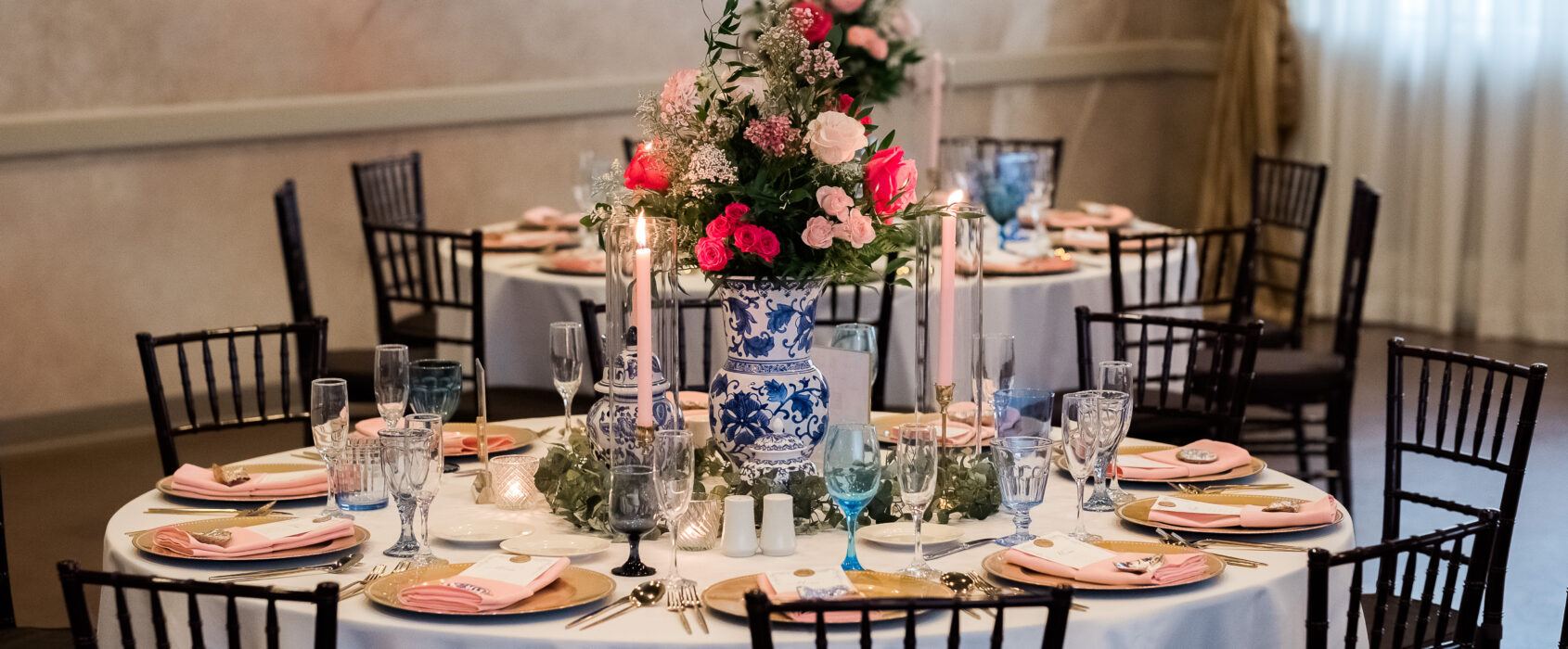 Elegant table setting with floral centerpiece, blue and white vases, candles, and pink napkins on white tablecloths.