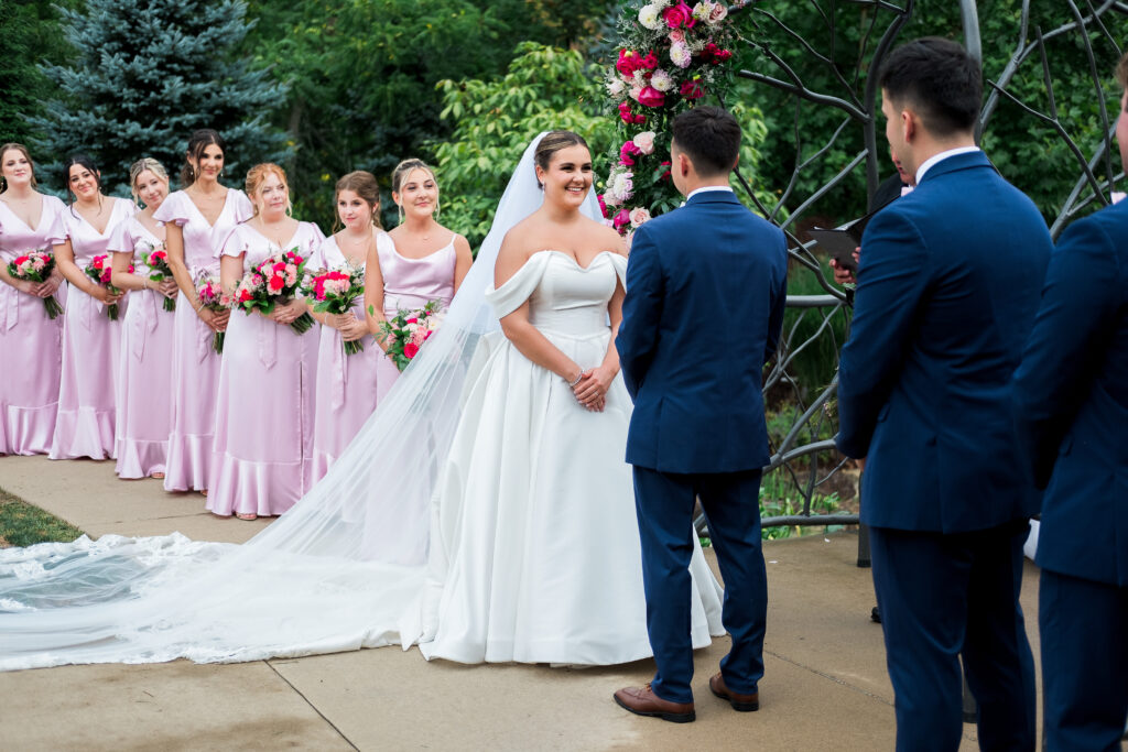 Bride and groom at an outdoor wedding ceremony, bridesmaids in pink dresses holding bouquets in the background.