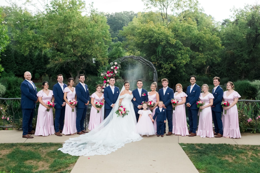 Wedding party posing outdoors; bride in white, bridesmaids in pink, groomsmen in blue suits holding bouquets.