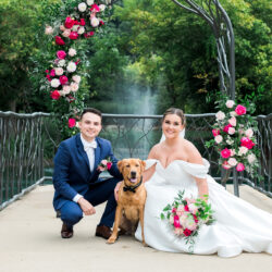 Bride and groom kneeling with dog under floral arch on a bridge, bride holding bouquet, fountain in background.