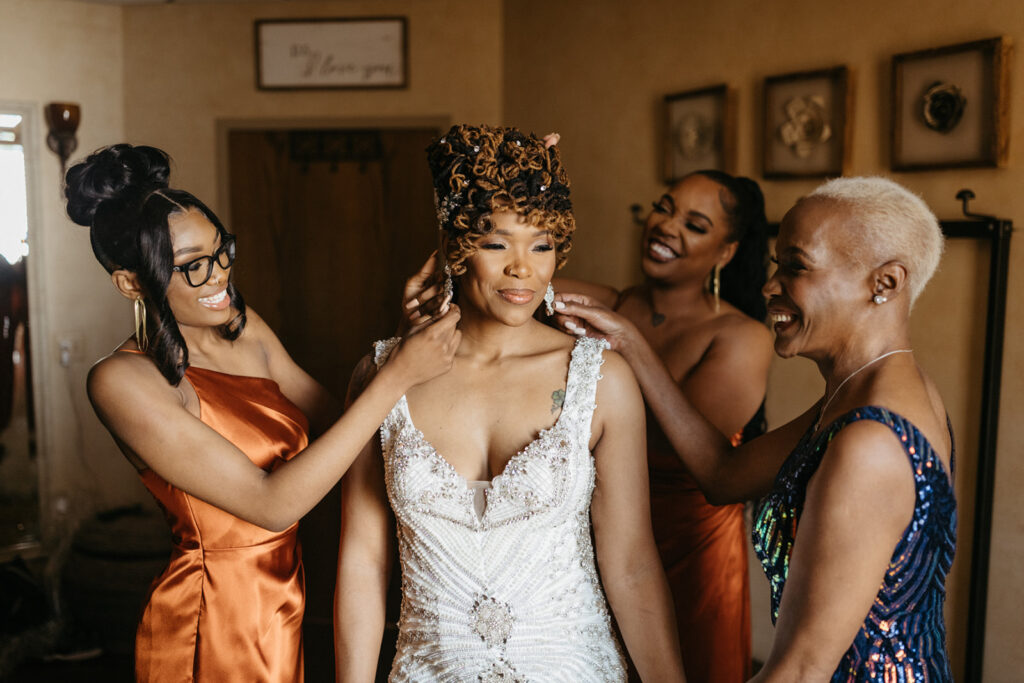 Bride in a sparkling dress is helped by three women, all smiling, in a warmly lit room.
