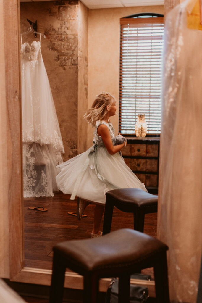 A young girl twirls in a dress, reflected in a mirror. A lacy wedding gown hangs nearby in a warmly lit room.