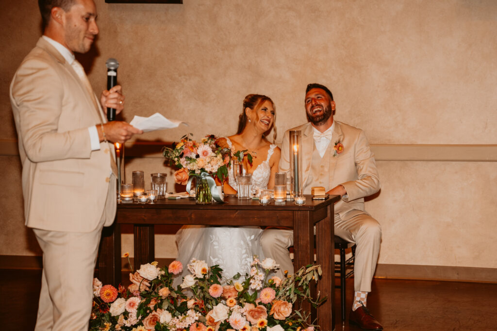 A couple in wedding attire laughs while seated at a table with flowers, as a man in a suit speaks into a microphone.