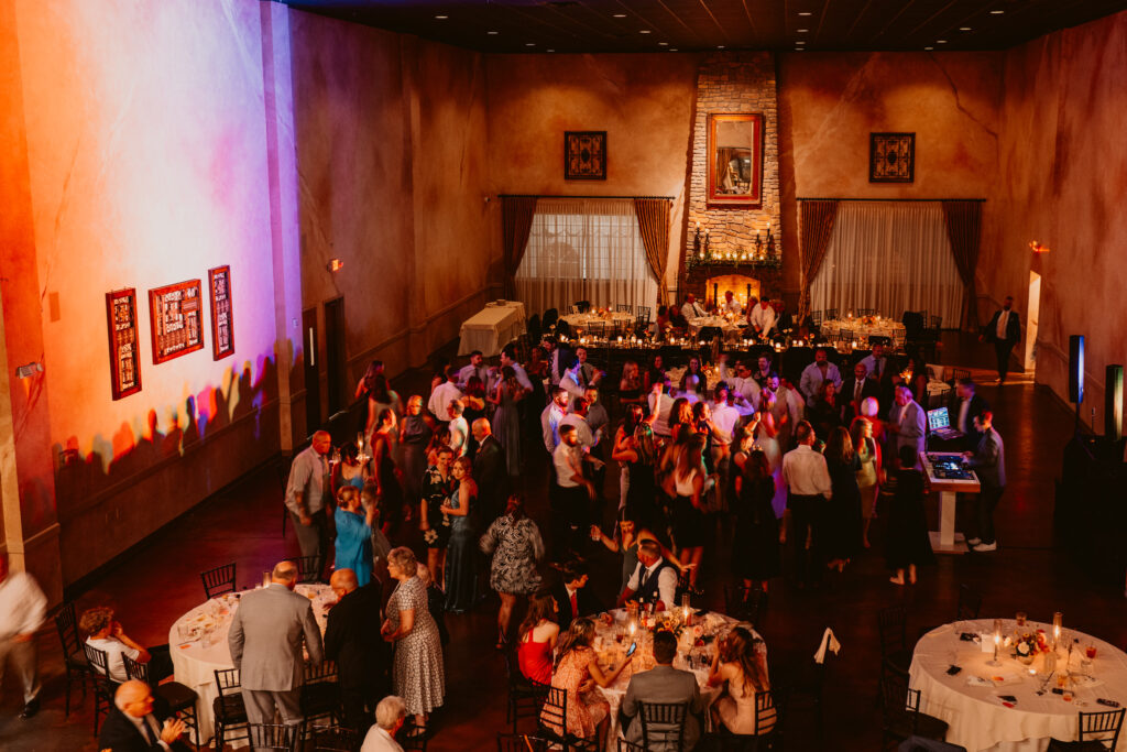 People dancing and socializing in a warmly lit banquet hall with tables and a DJ booth.