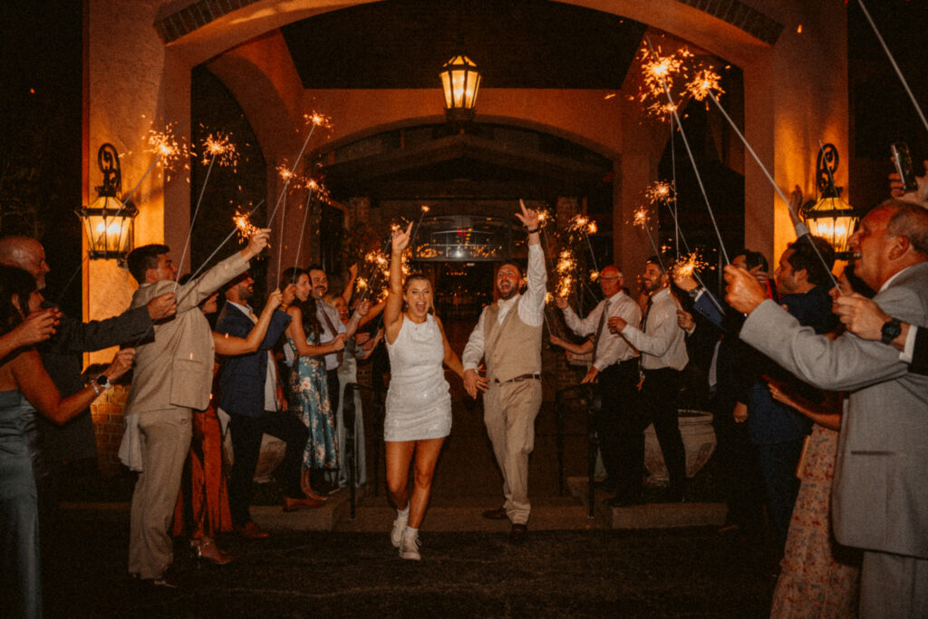 A couple joyfully exits a party under lit sparklers, surrounded by cheering guests in an evening setting.