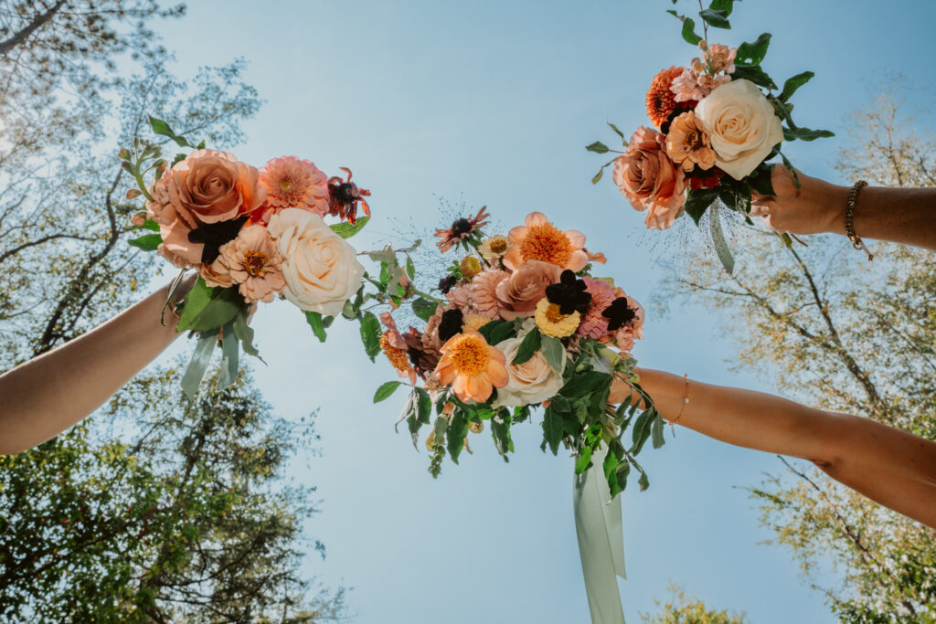 Three hands holding bouquets of roses and flowers overhead against a clear blue sky.