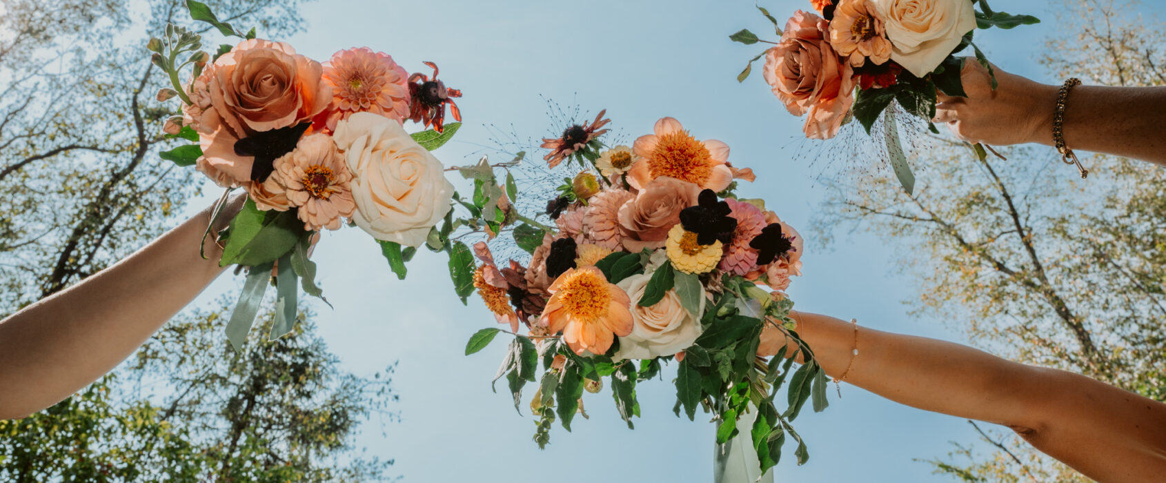 Three hands holding bouquets of roses and flowers overhead against a clear blue sky.