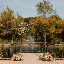 Outdoor wedding altar with pink and white flowers, set on a bridge with a fountain and trees in the background.