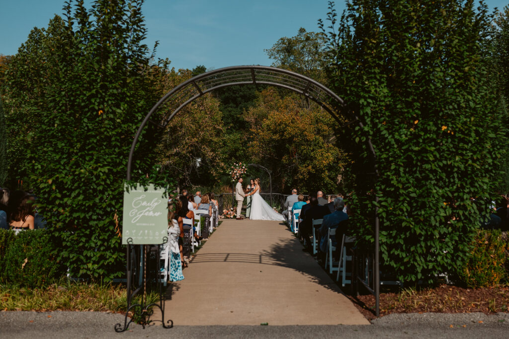 Outdoor wedding ceremony with a couple at the altar, guests seated, and a sign on the left under an archway.