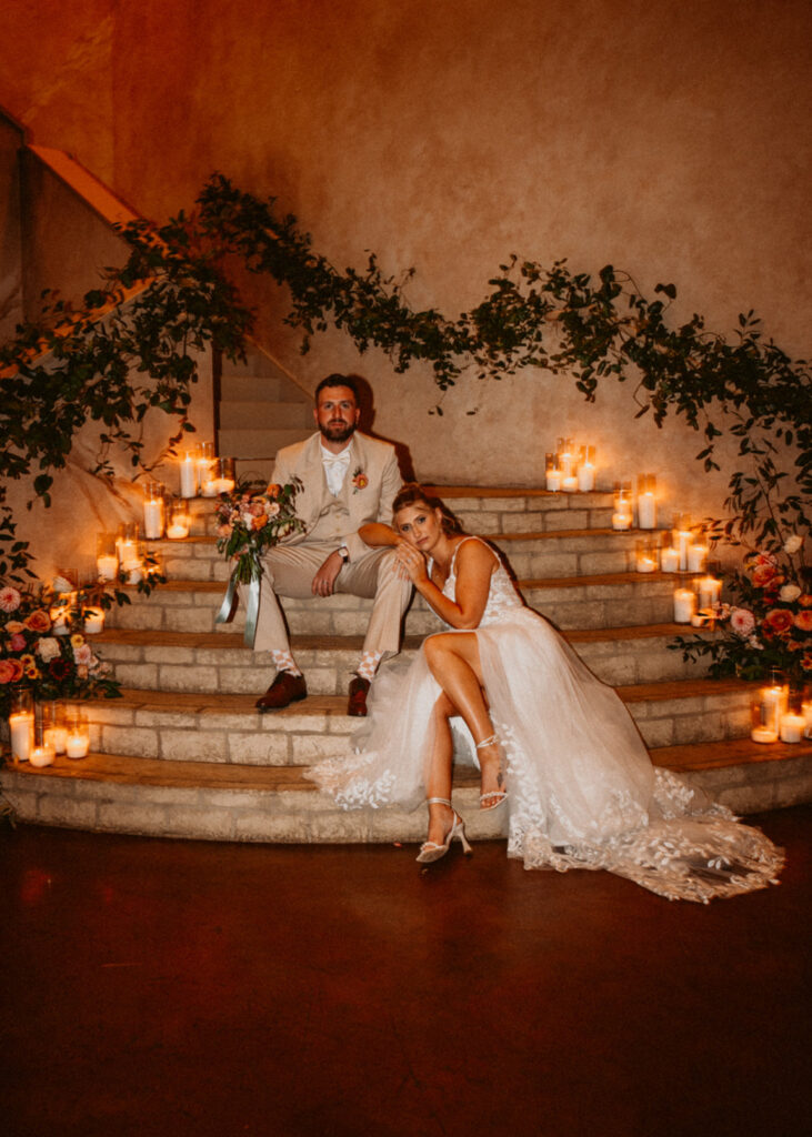 Bride and groom sitting on candle-lit steps, surrounded by greenery and flowers.