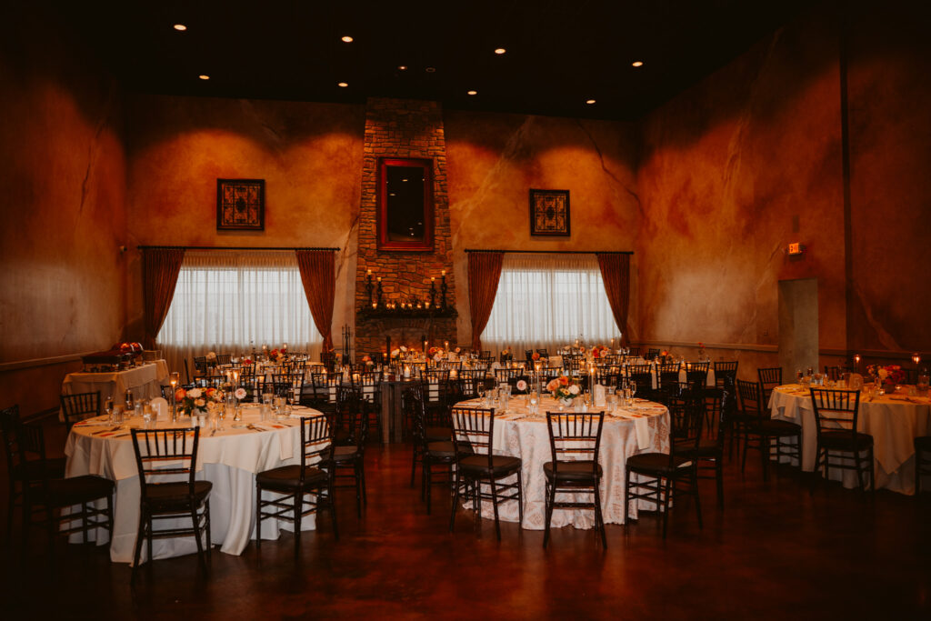 Dimly lit banquet hall with round tables set for a formal event, featuring draped curtains and a stone fireplace.