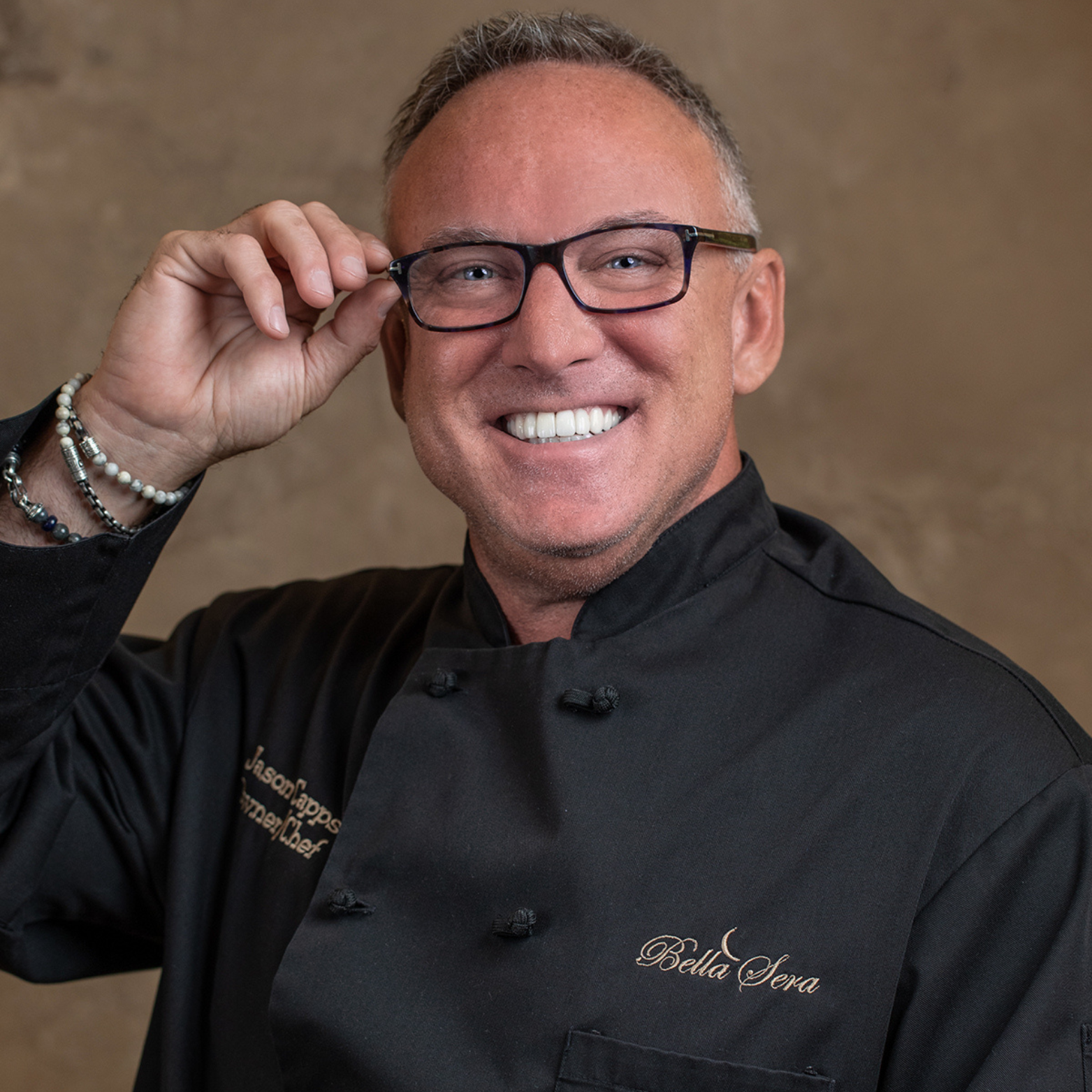 Smiling man wearing glasses and a black chef's coat with "Bella Sera" embroidered, adjusting his glasses.