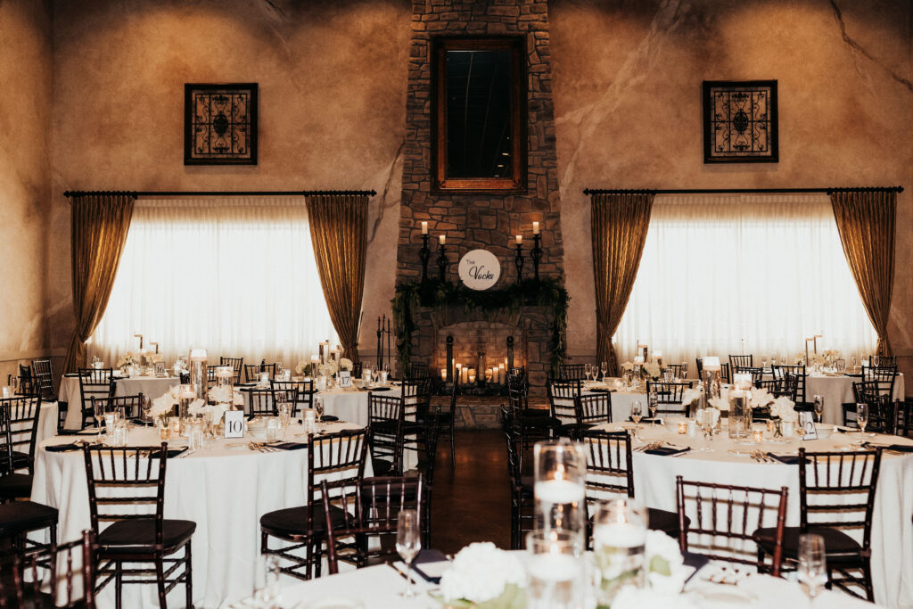 Elegant wedding reception venue with round tables, white linens, and candles, centered around a stone fireplace.