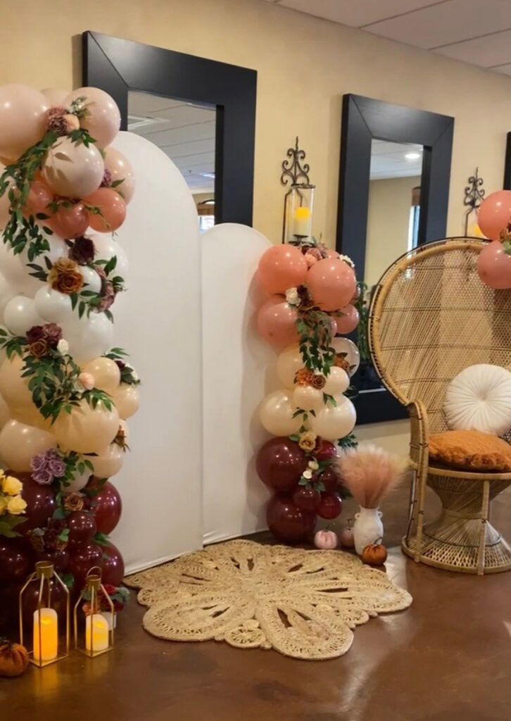 Decorative event setup with balloon arches, wicker chair, candles, and flowers in neutral tones.