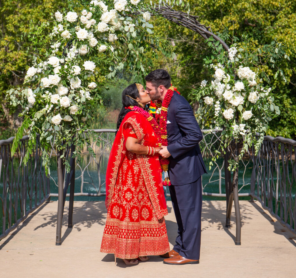 Couple in traditional attire kissing under a floral arch at an outdoor wedding ceremony.