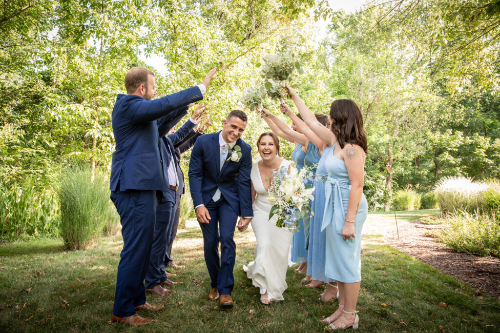 Bride and groom smiling under a human arch made by the bridal party in a sunny, green garden setting.