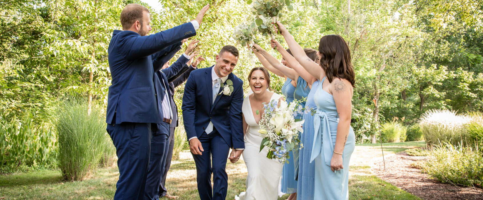 Bride and groom smiling under a human arch made by the bridal party in a sunny, green garden setting.