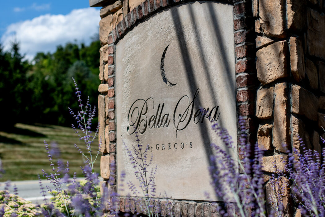 Stone sign reading "Bella Sera" with lavender flowers in the foreground and a blue sky background.